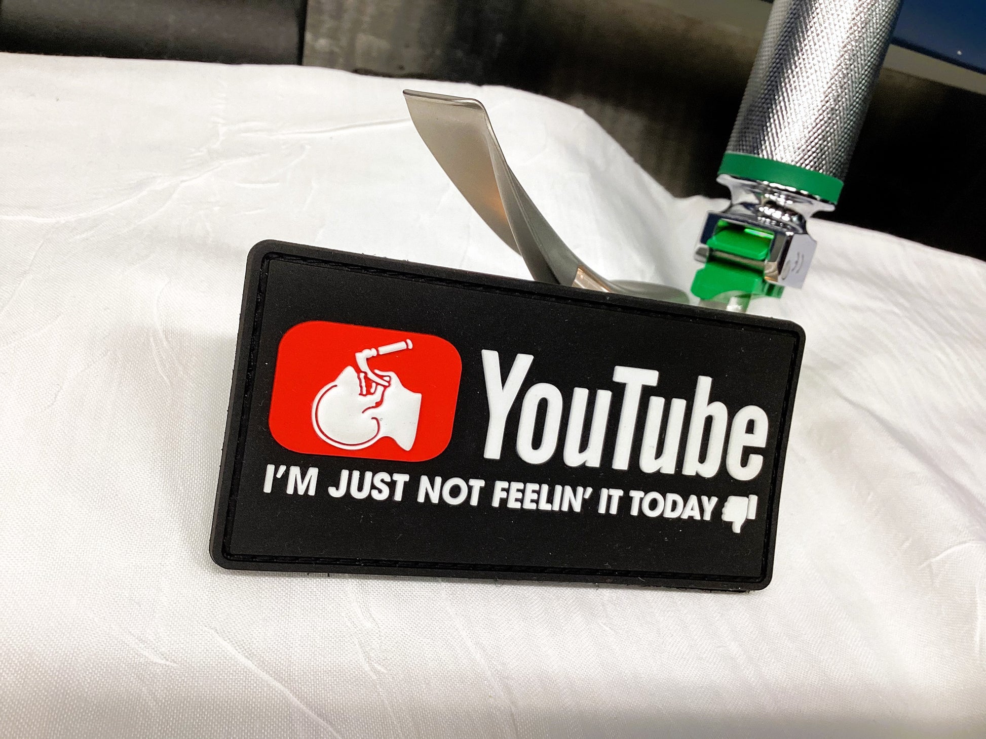 YouTube Intubation patch from skell city, leaning against a laryngoscope blade against a white hospital sheet background.