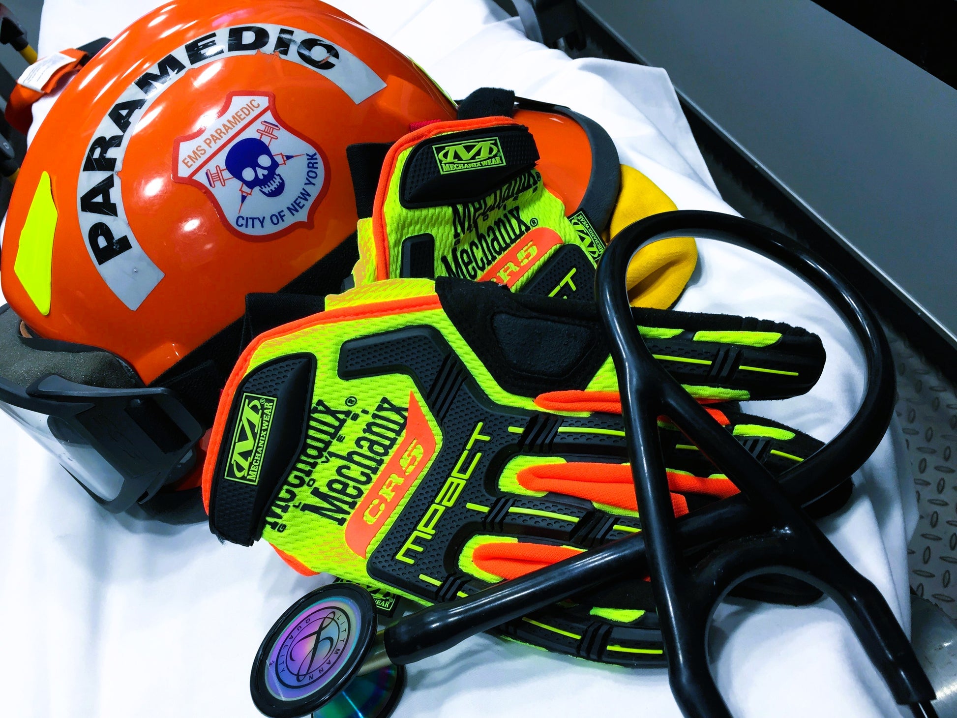 NYC skell city EMS Paramedic patch reflective sticker on an orange ems bunker gear helmet with extrication gloves and a Littman cardiology 3 rainbow stethoscope on a stryker stretcher.