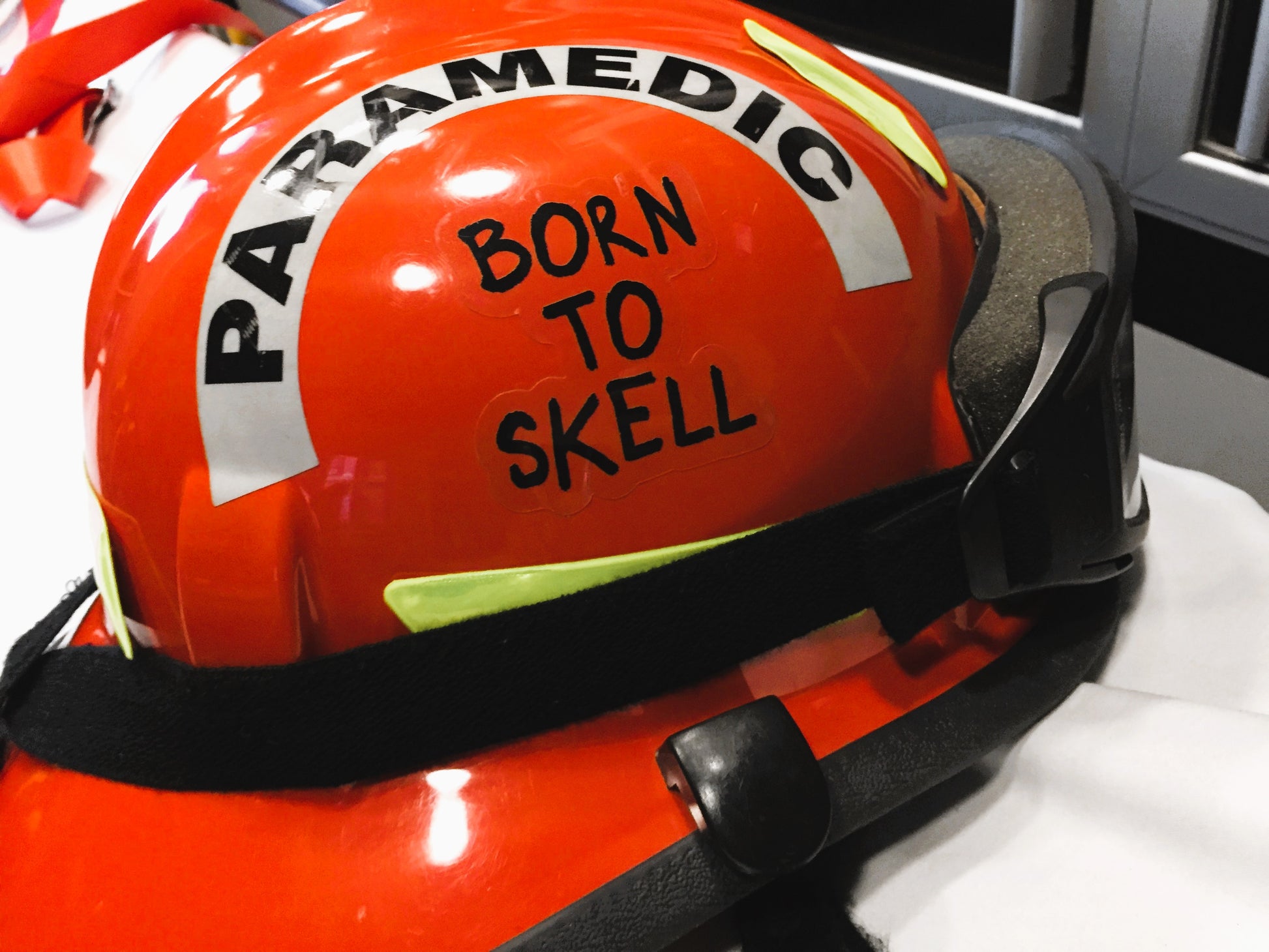 Paramedic Orange Bunker Gear Helmet On a Stretcher, which has a Clear Born To Skell Sticker On the Side of the helmet.