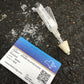 Skell City Medicaid card, key to the city, on the concrete street, next to crushed Luer-Jet and broken narcan naloxone vial inside. 