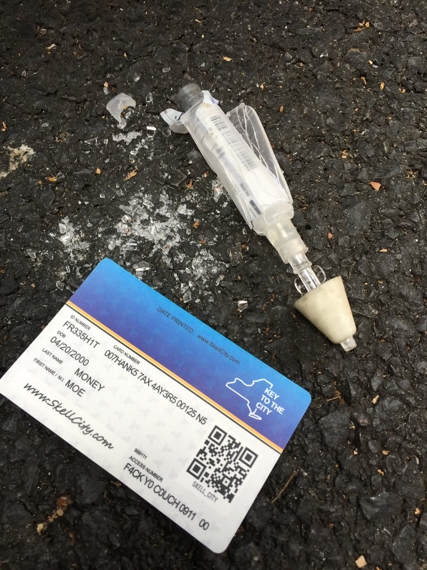 Skell City Medicaid card, key to the city, on the concrete street, next to crushed Luer-Jet and broken narcan naloxone vial inside. 