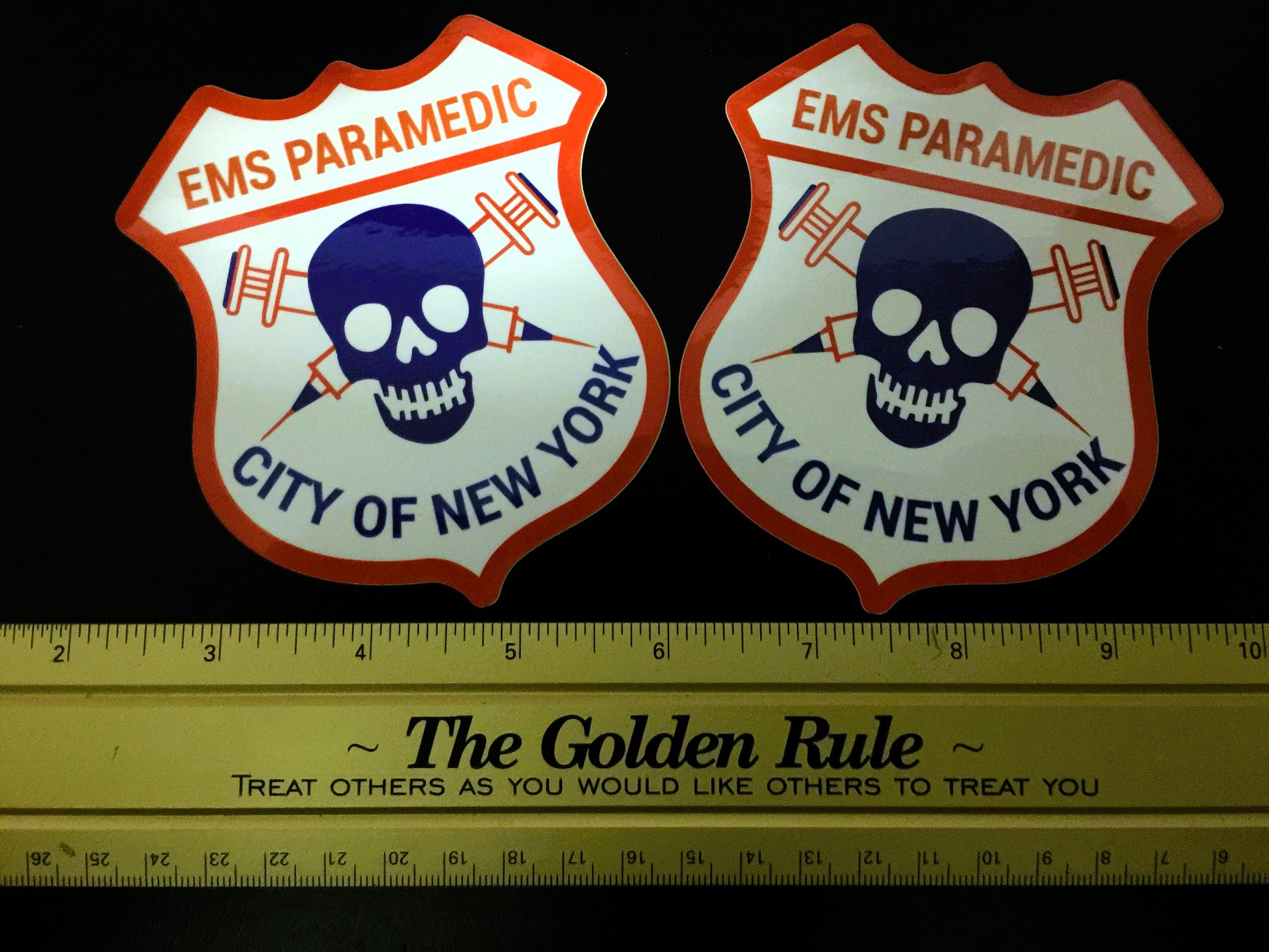 The NYC EMS Paramedic Patch Glossy Stickers against a black background, next to a golden ruler with the golden rule.