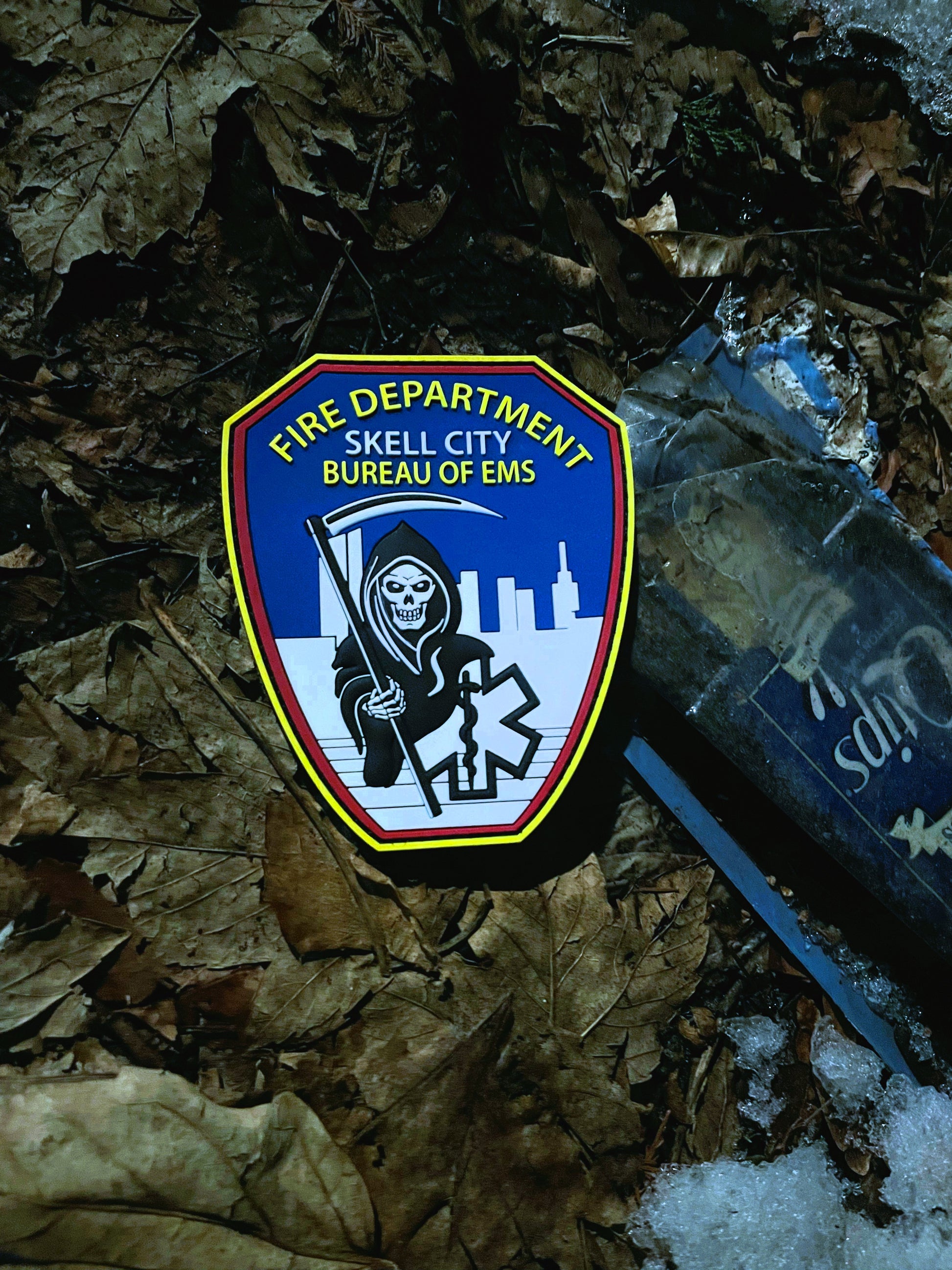 EMS patch from skell city lying on dirty wet leaf covered ground, next to garbage.